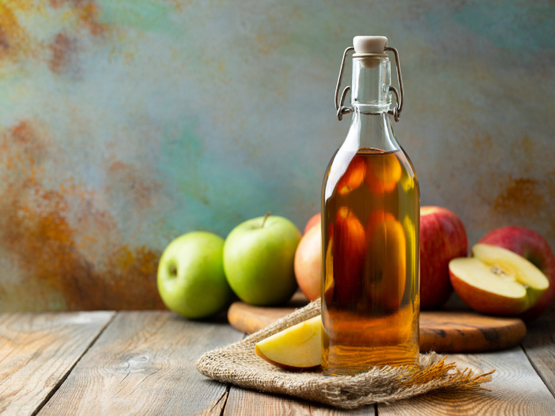 Photo of a bottle of applie cider vinegar with green and red apples in the background
