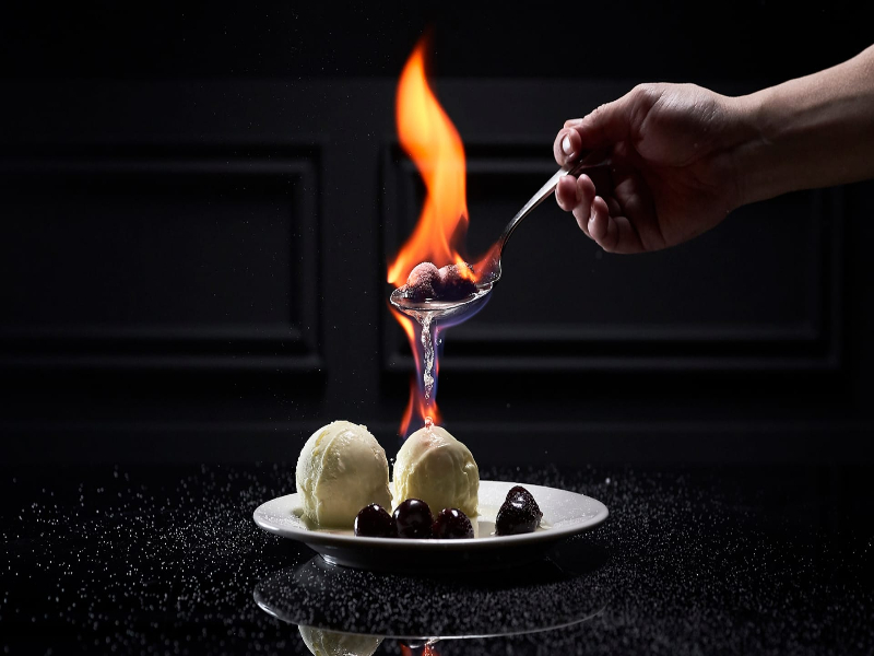 A photo of a hand holding a flaming cherry being drizzled over 2 scoops of vanilla ice cream