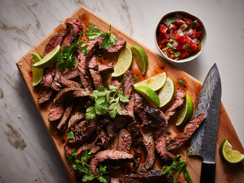 A photo of carne asada sliced on a cutting board with limes and salsa