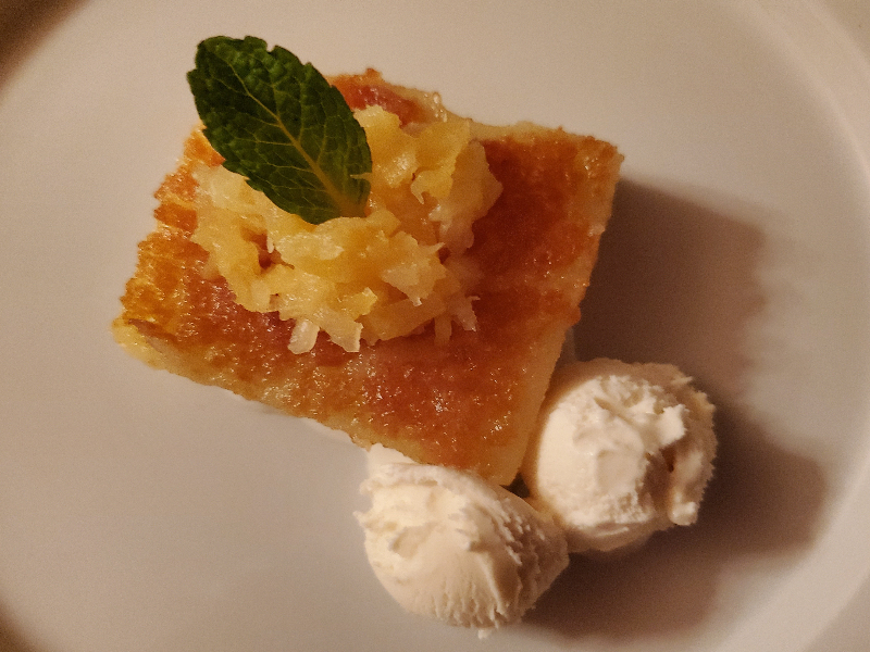 A photo of mochi cake topped with crushed pineapple and a mint left with two small scoops of vanilla ice cream on the plate.