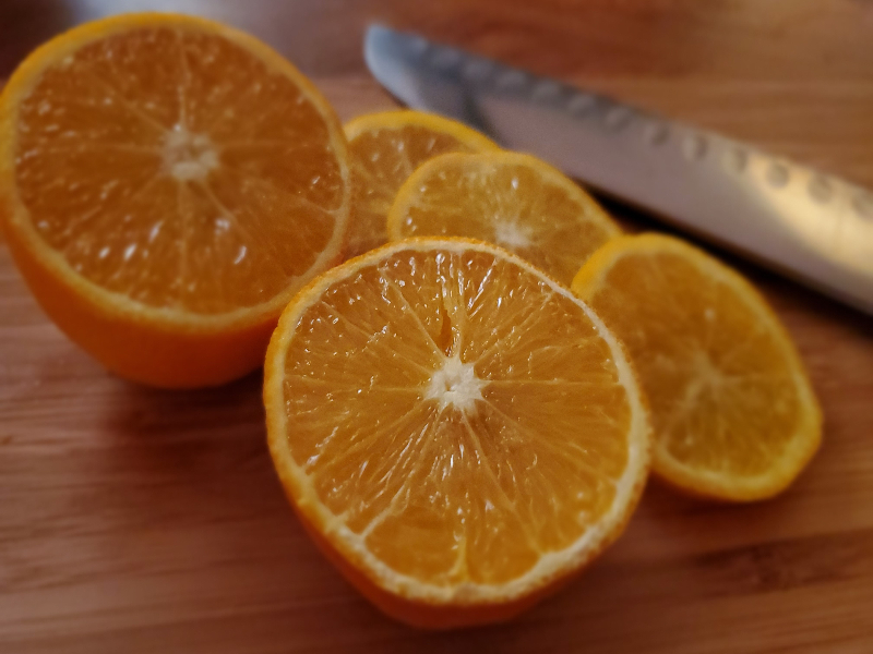 A photo of an orange cut in half with slices and a knife on a wooden board.