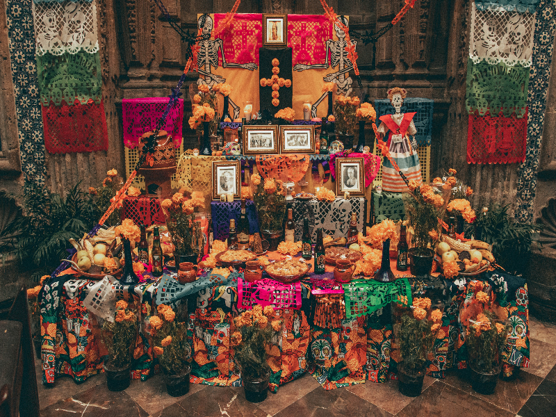 A photo of an altar celebrating Dia de los Muertos with flowers, photos, candles, food, and drink.