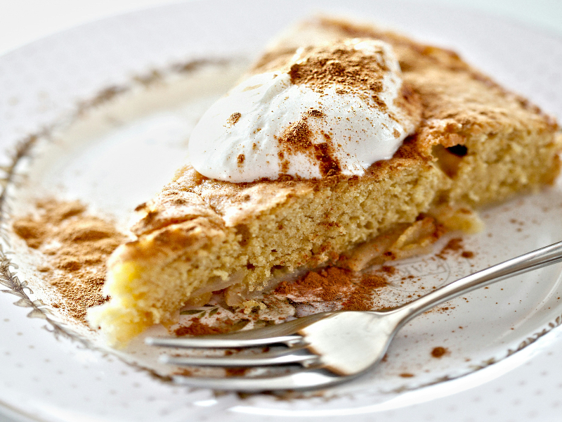 Photo of a slice of apple cake on a glass plate with a fork and topped with a dollop of whipped cream and cinnamon.