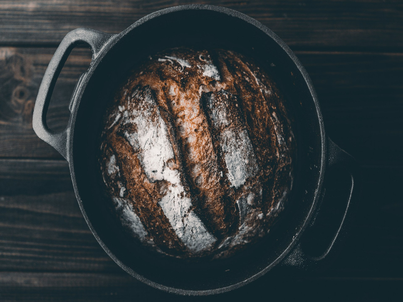 A photo of a baked round loaf of mesquite country bread inside a Dutch oven resting on a wood table.